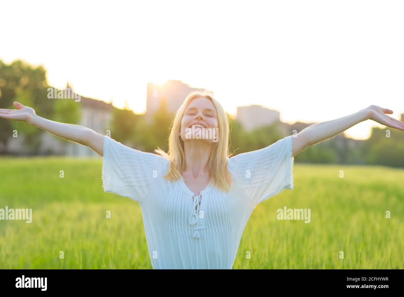 Happy young beautiful woman with arms open in the air in a green environment during sunset. Well-being and freedom. Stock Photo