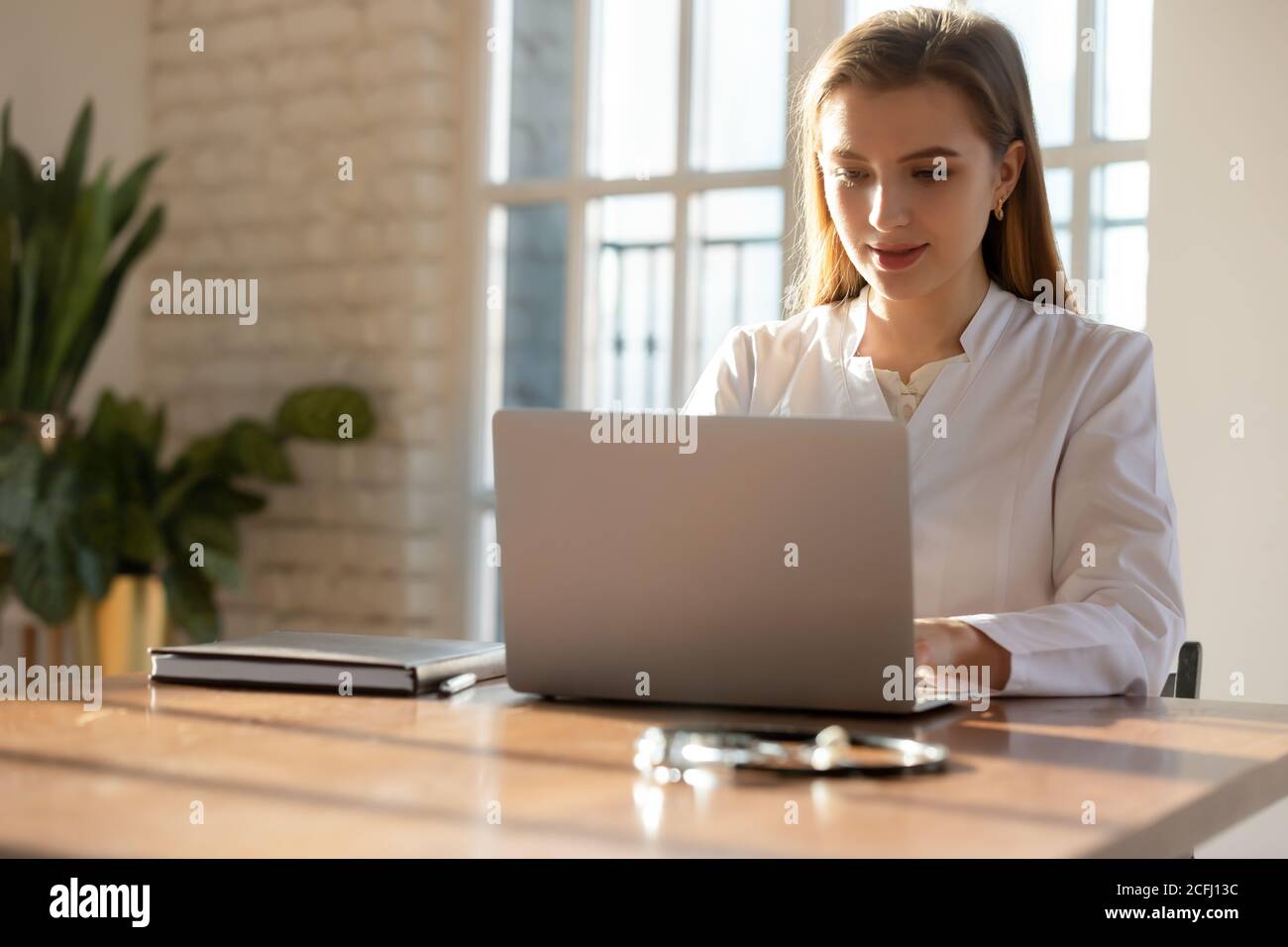 Focused young woman doctor working on laptop online in office Stock Photo