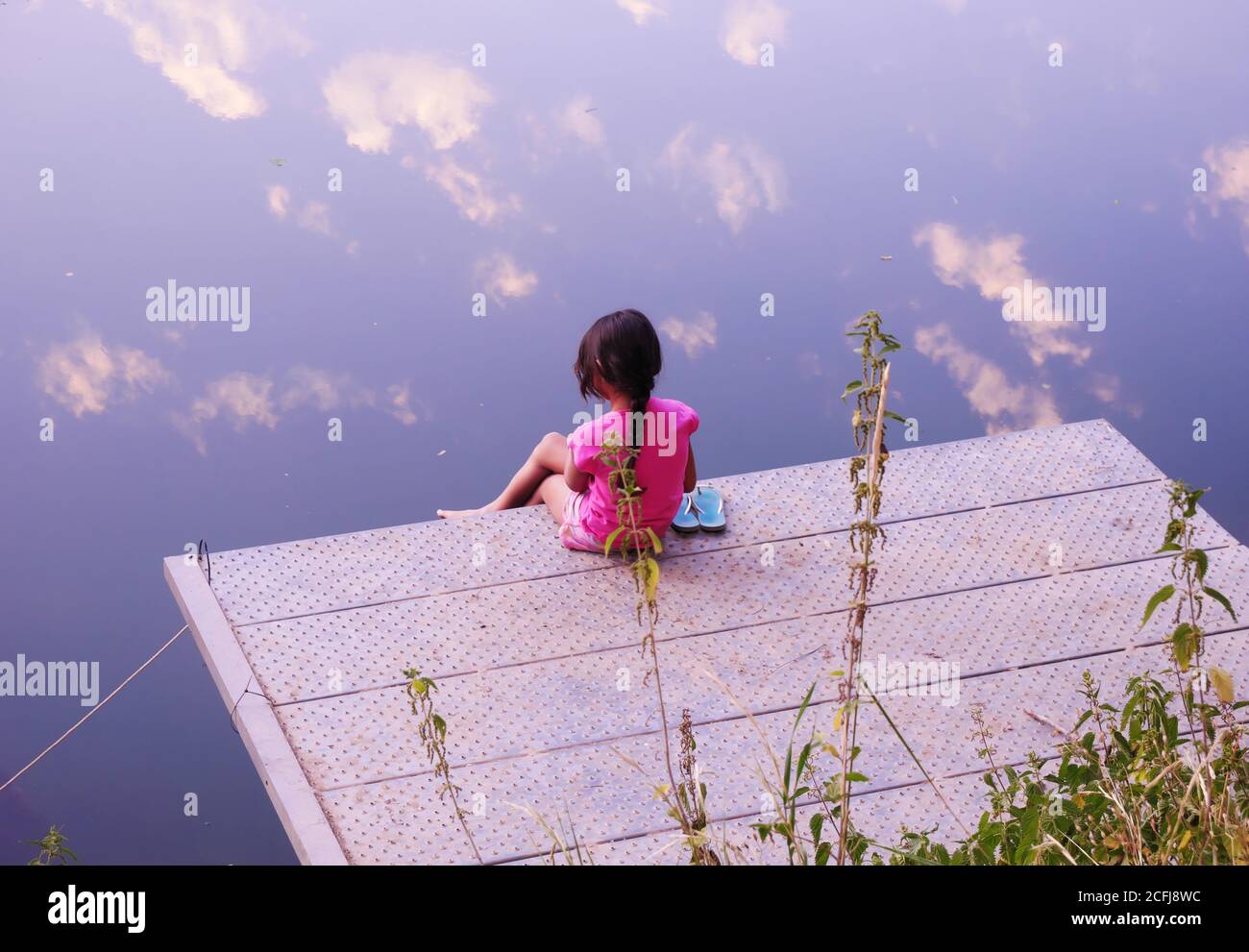 Little girl seen from behind, sitting on a pontoon and contemplating the clouds that reflect in the river during a sunset Stock Photo