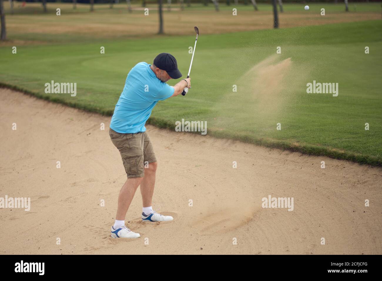 Golfer hitting his ball clear of the bunker or sand hazard in a puff of flying sand as he plays the shot onto the fairway Stock Photo