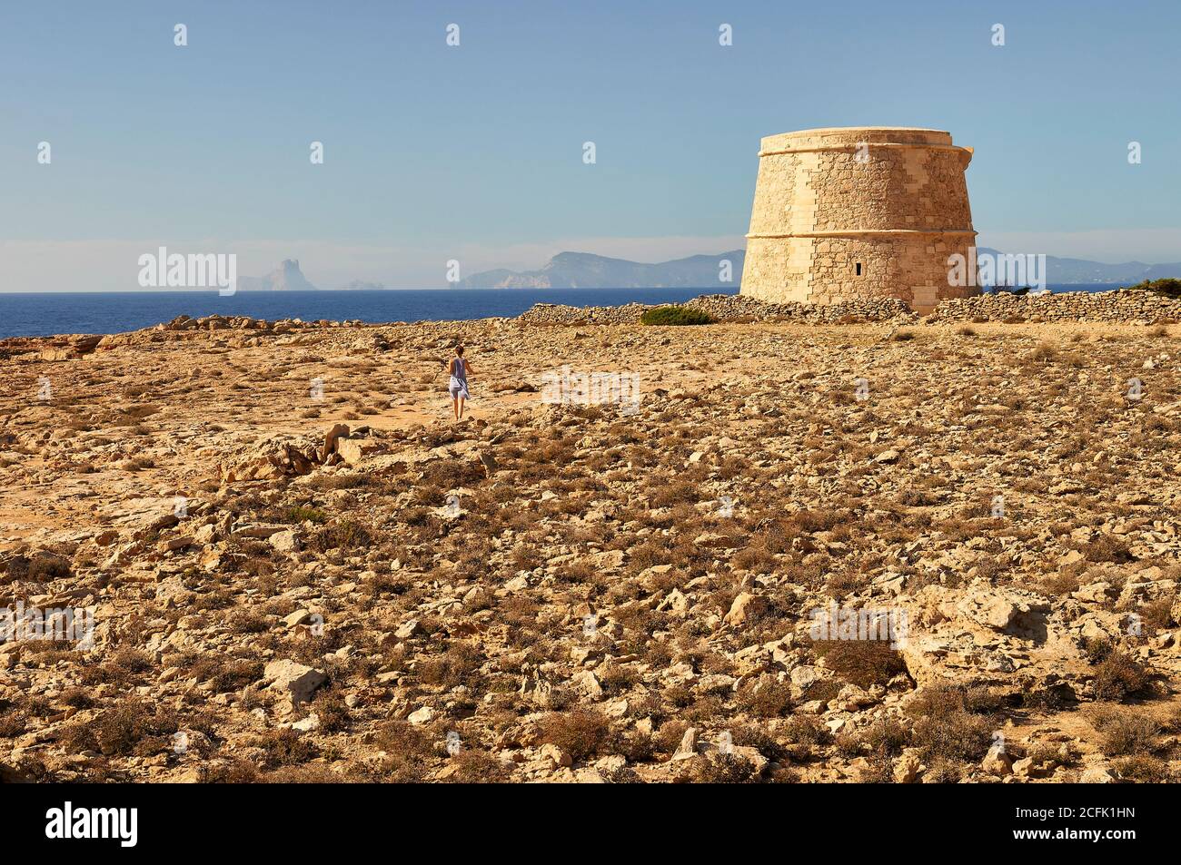Woman walking through a small trail towards the Sa Gavina defense tower with Es Vedrá island in the distance (Formentera, Balearic Islands, Spain) Stock Photo
