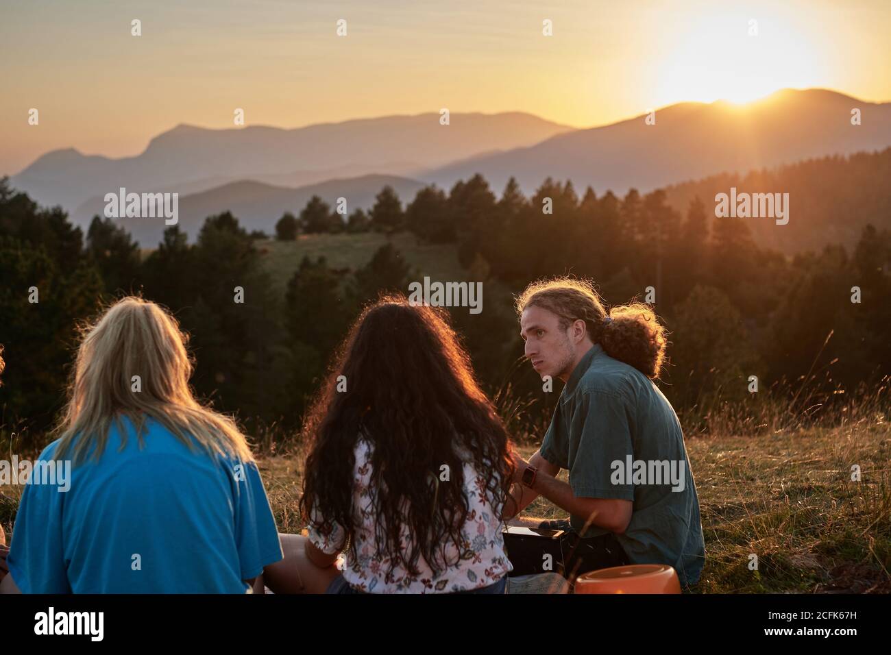 Back view of group of traveling friends relaxing on grass near camping tent and enjoying summer vacation during sundown in highland area Stock Photo