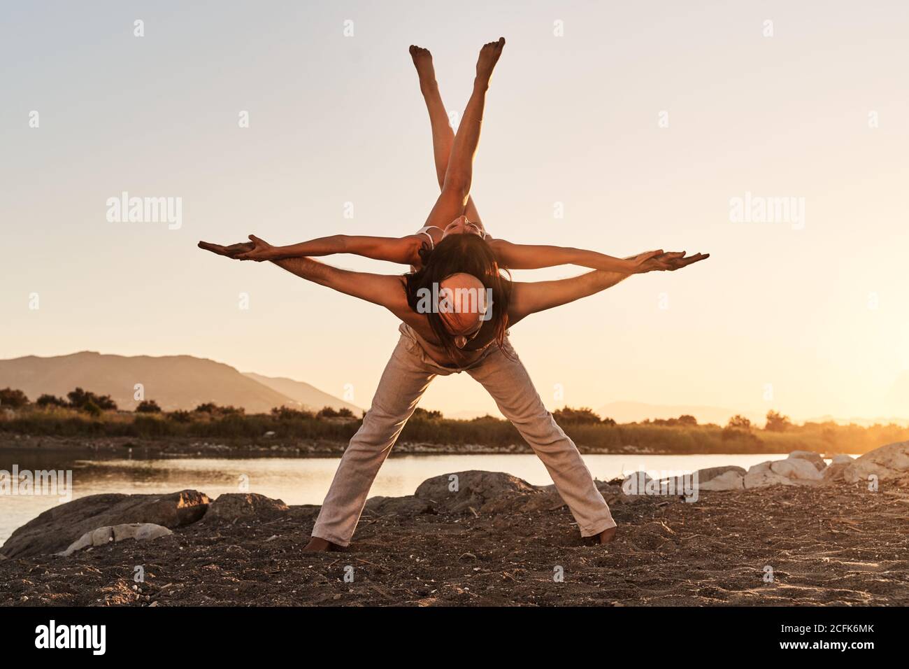 Strong man with slender woman on back balancing with raised crossed legs while practicing acro yoga during sunset in summer Stock Photo