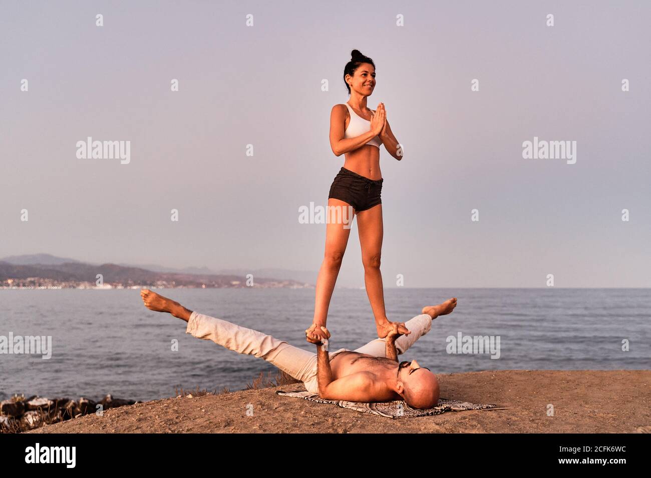 Slim woman in Mountain pose with Namaste gesture balancing on hands of strong man while doing acroyoga at seaside Stock Photo