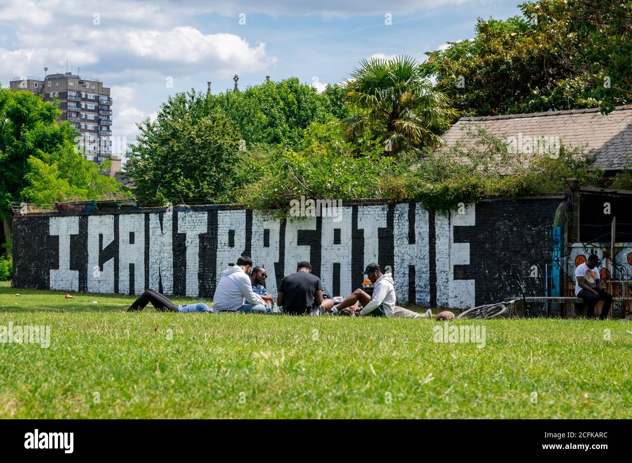 A group of young people relaxing in a park by Brick Lane with a view of a brick wall with the words i cant breath . Stock Photo