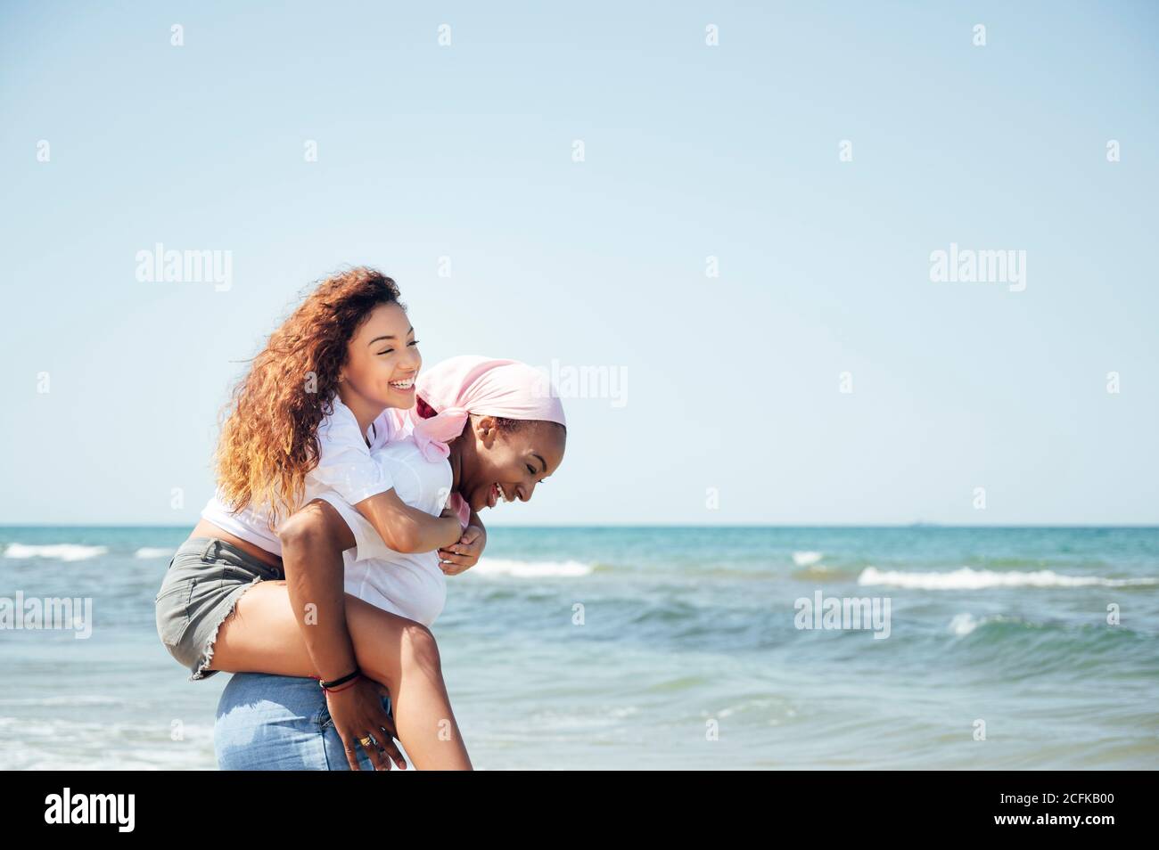Delighted black mother piggybacking young daughter while having fun with closed eyes at seaside in summer Stock Photo