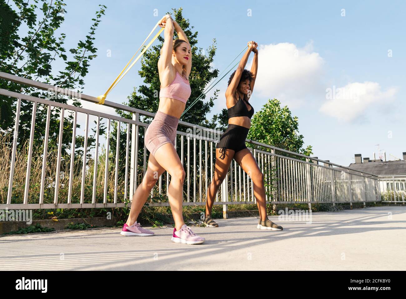 Low angle of diverse female athletes in sportswear doing exercises with resistance bands near metal railing on terrace in summer Stock Photo