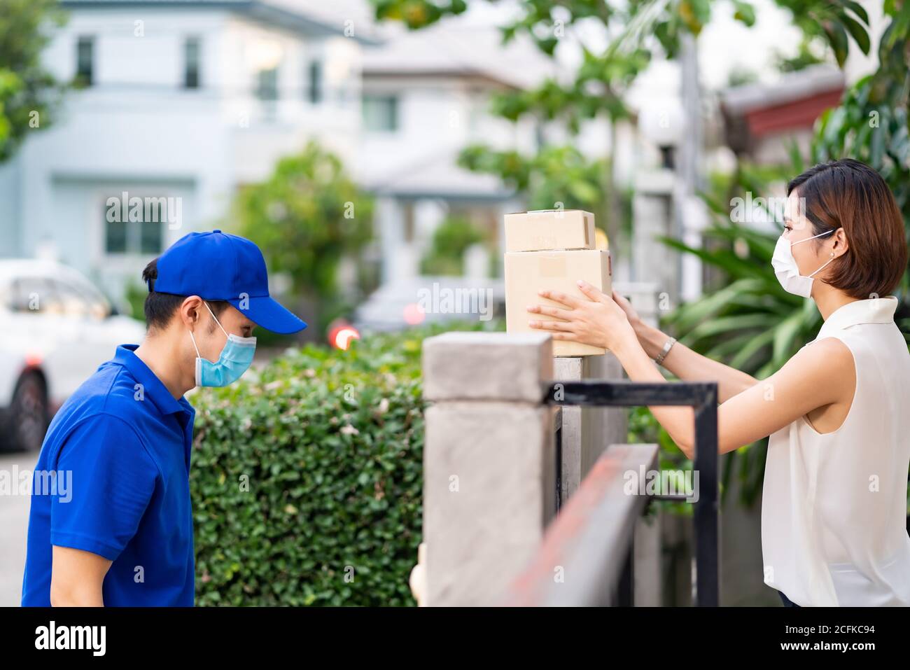 Side view asian woman with face mask customer take shopping packages from the fence post that delivery man put them on for contactless delivery. This Stock Photo