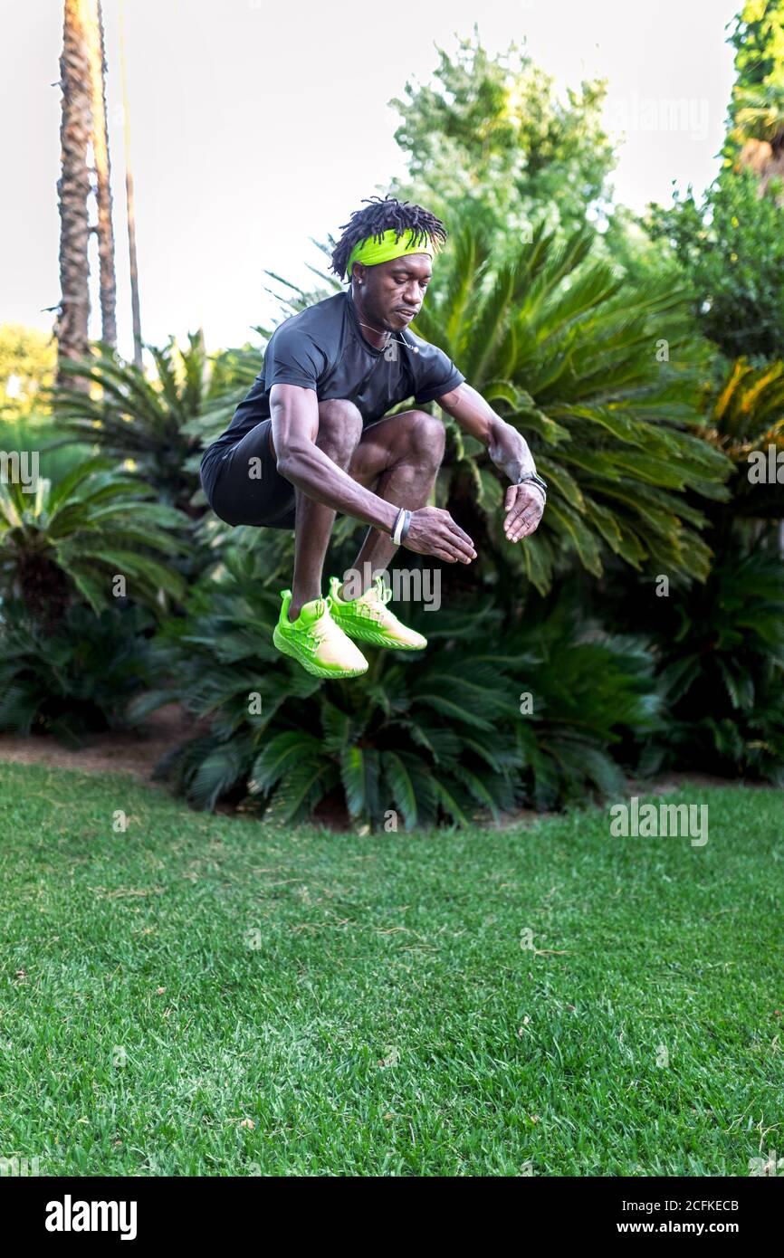 Full body of young African American male athlete jumping high above green lawn while working out in tropical park Stock Photo