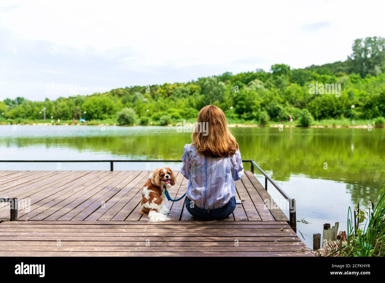 Rear view shot of woman wearing casual clothes while sitting on the pier next to her puppy. Copy space. Stock Photo
