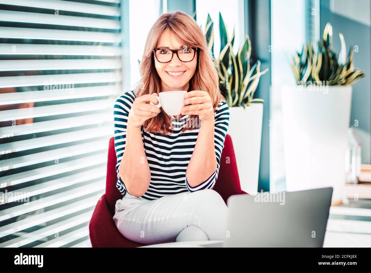 Shot of happy woman sitting in the armchair and drinkig her morning coffee while working from home. Home office. Stock Photo