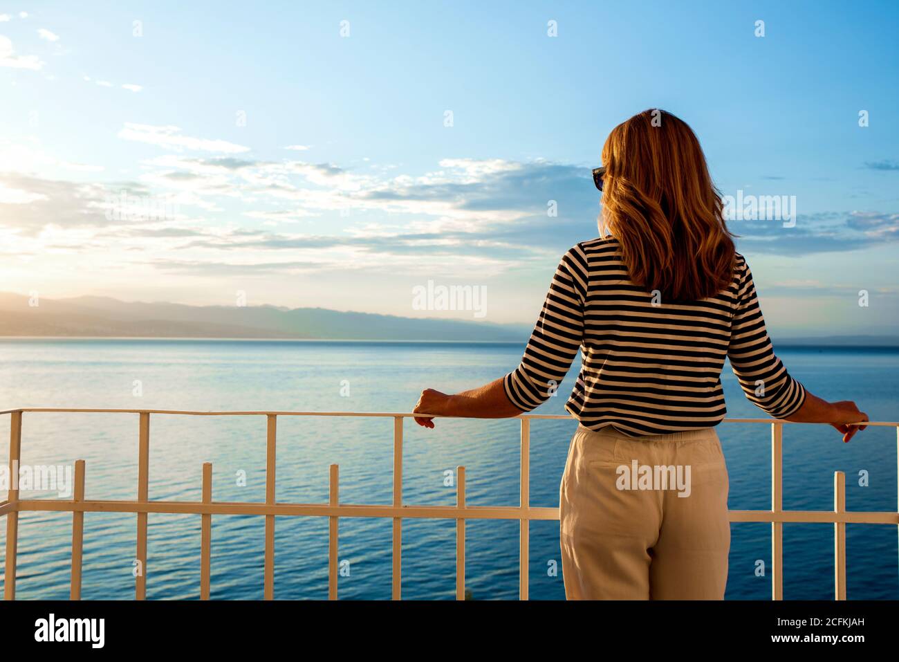 Shot of pretty woman wearing straw hat and sunglasses while standing on balcony and looking at sea view. Daydreaming. Stock Photo
