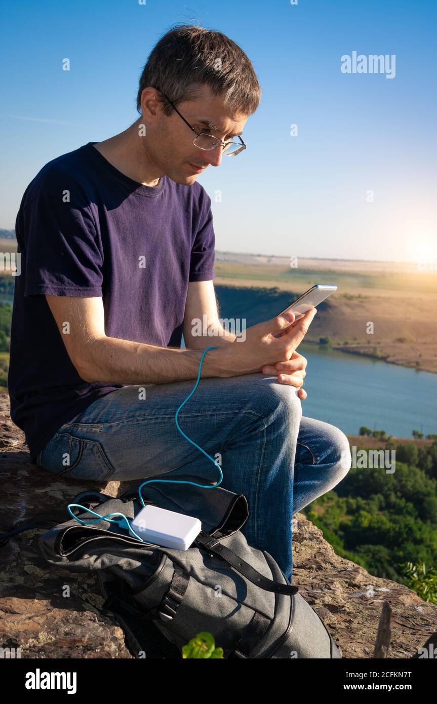 Man on a hike uses smartphone while charging from the power bank on the rock at dawn. Stock Photo