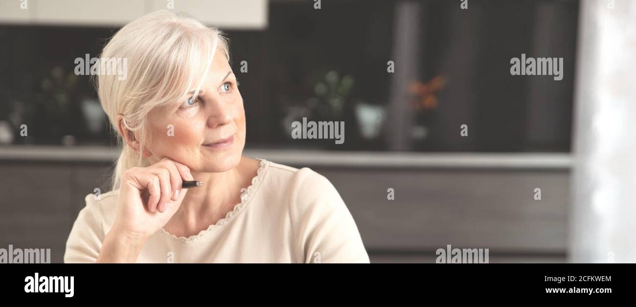Pensioner woman holding a pen, sits at home and looks at the window. Cheerful senior woman portrait. Concept of a happy retirement Stock Photo