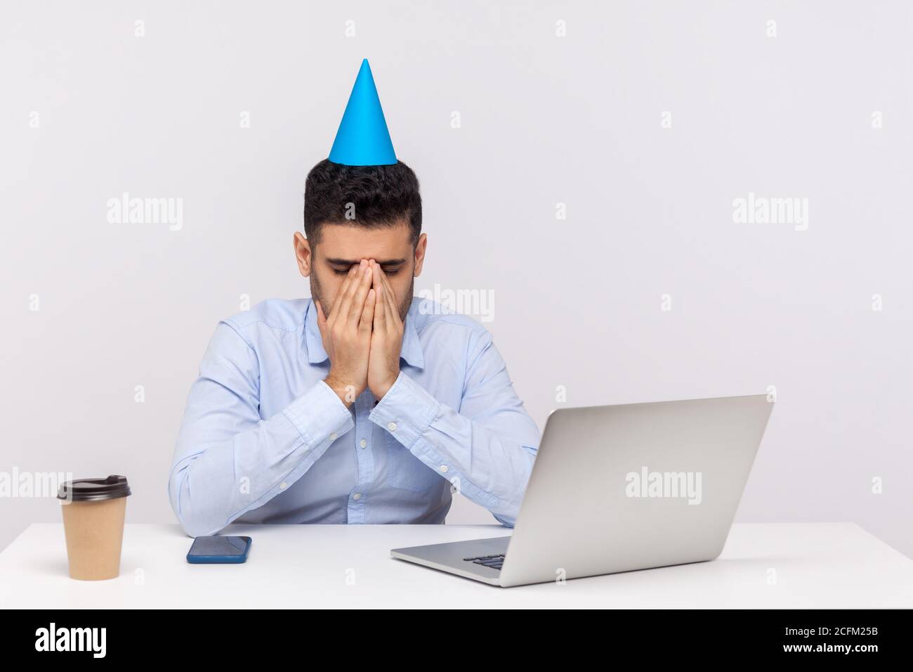 Upset depressed employee guy with party cone on head crying at workplace, hiding face feeling desperate lonely on birthday has to stay in office. indo Stock Photo