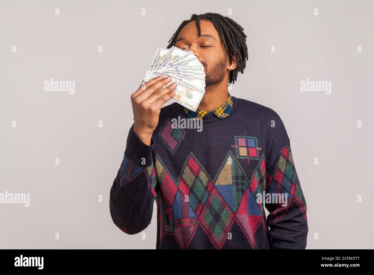 Happy satisfied african guy with dreadlocks and beard sniffing big batch of money enjoying big salary. Indoor studio shot isolated on gray background Stock Photo