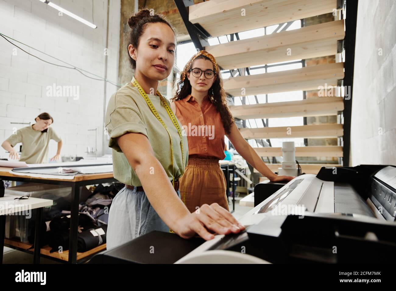 Two young tailors standing by large industrial printer under staircase in studio Stock Photo