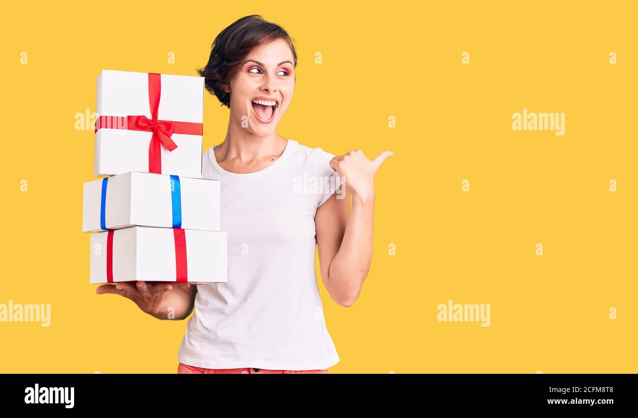 Beautiful young woman with short hair holding gifts pointing thumb up ...