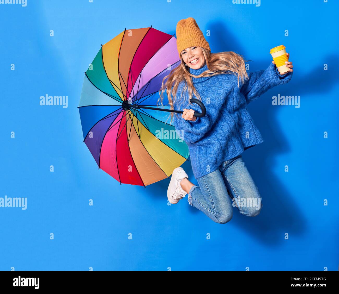 Young beautiful woman wearing winter clothes smiling happy. Jumping with smile on face holding colorful umbrella drinking takeaway coffee over isolate Stock Photo