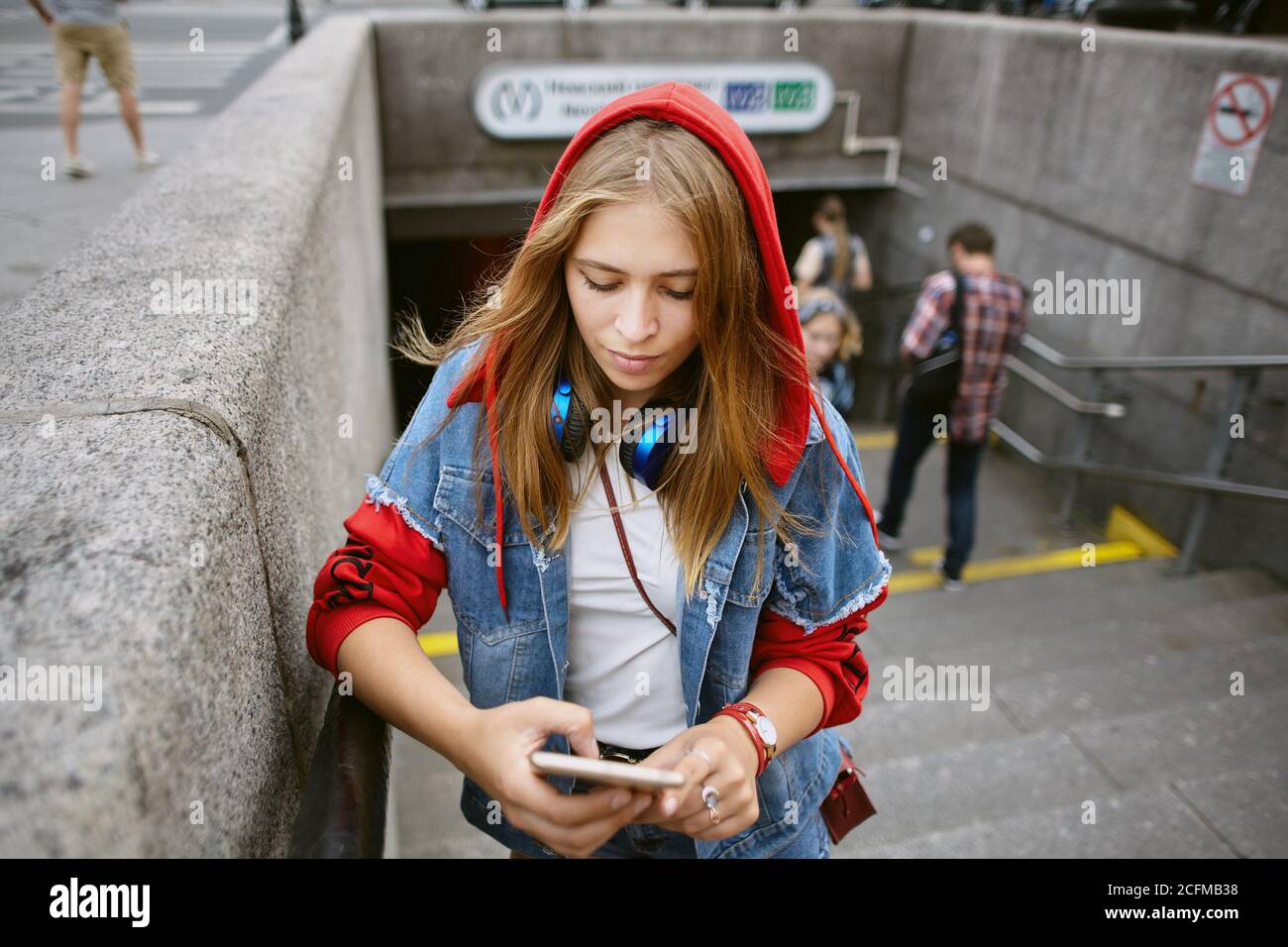 A young beautiful woman in red hoodie is standing at the entrance of the subway with  mobile phone in hands. Stock Photo