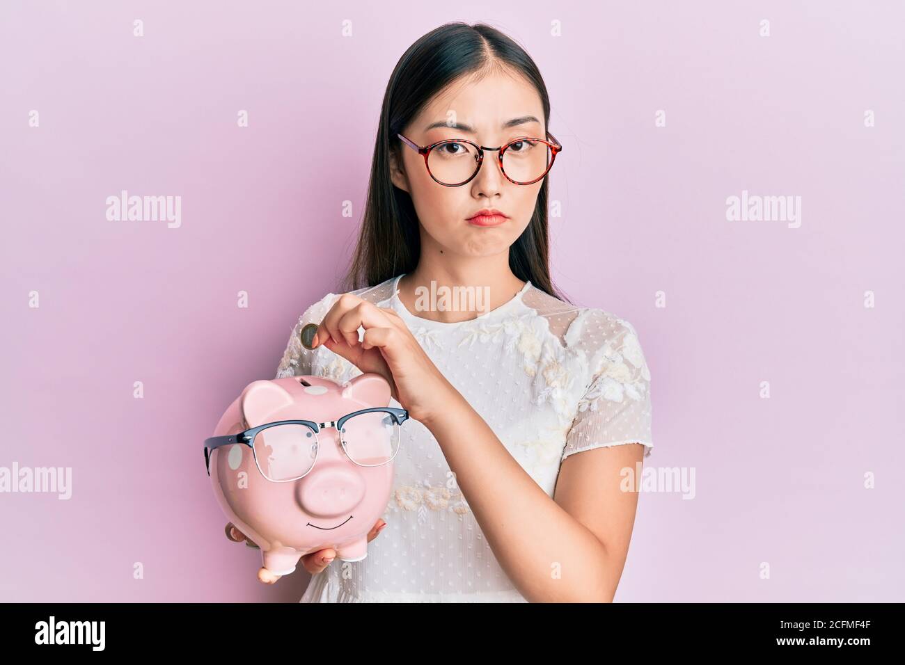 Young chinese woman holding piggy bank with glasses depressed and worry ...
