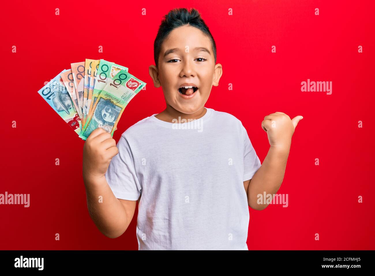 Little boy hispanic kid holding australian dollars pointing thumb up to the side smiling happy with open mouth Stock Photo