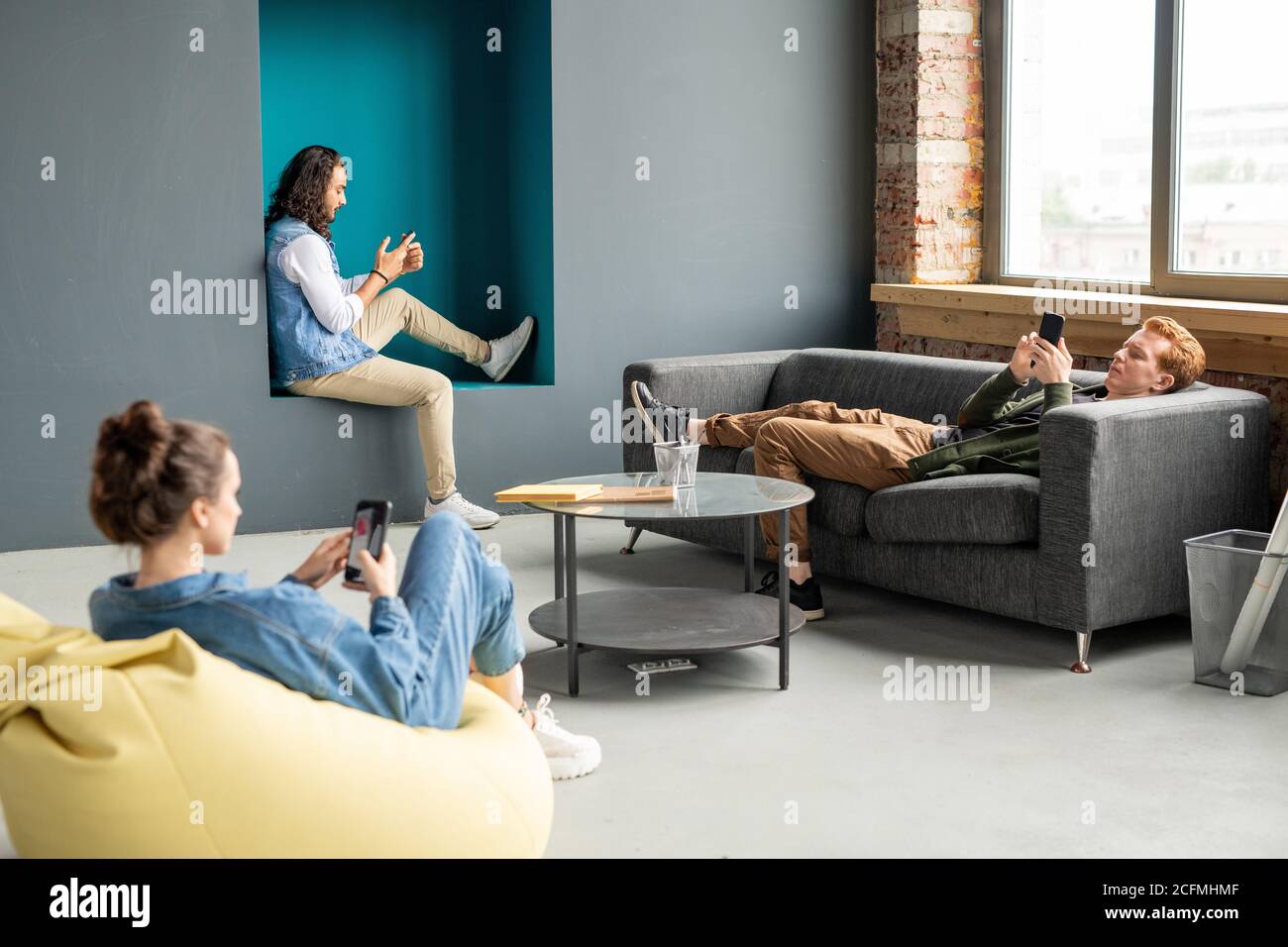 Three young restful specialists using mobile gadgets while relaxing in office Stock Photo