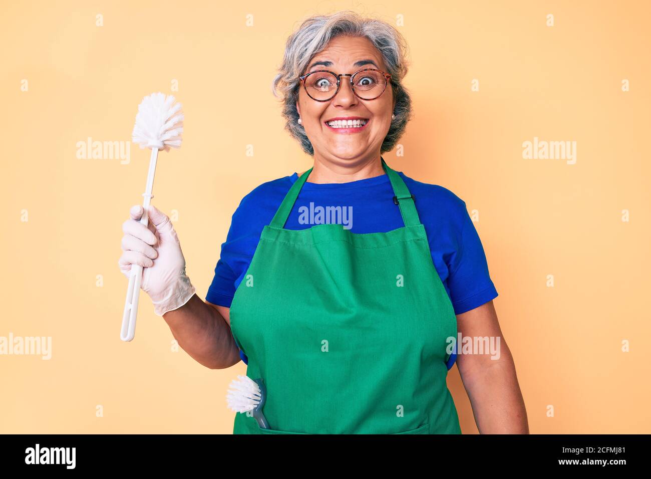 Senior hispanic woman wearing apron holding brush looking positive and ...