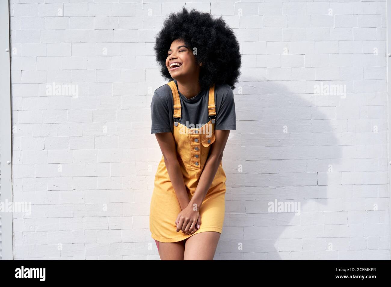 Happy African lady with Afro hair laughing standing against white brick wall. Stock Photo