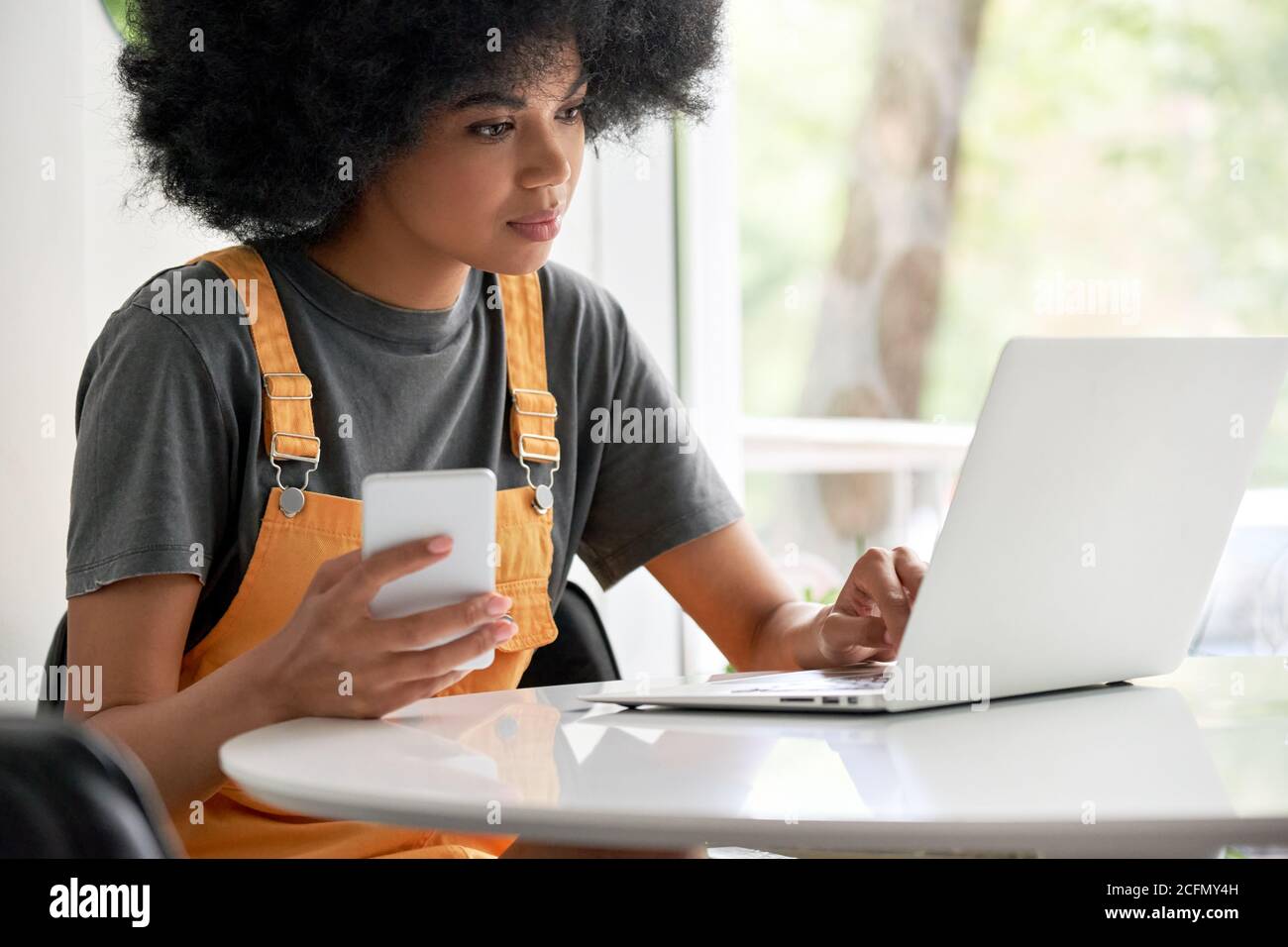 Young African woman student using laptop holding smartphone sitting at table. Stock Photo