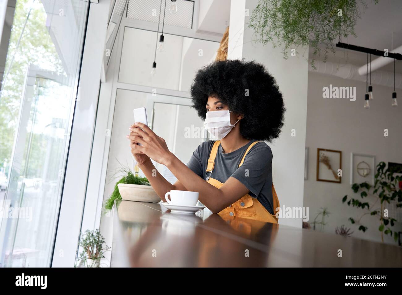 African lady wearing face mask using phone sit at table in modern cafe. Stock Photo