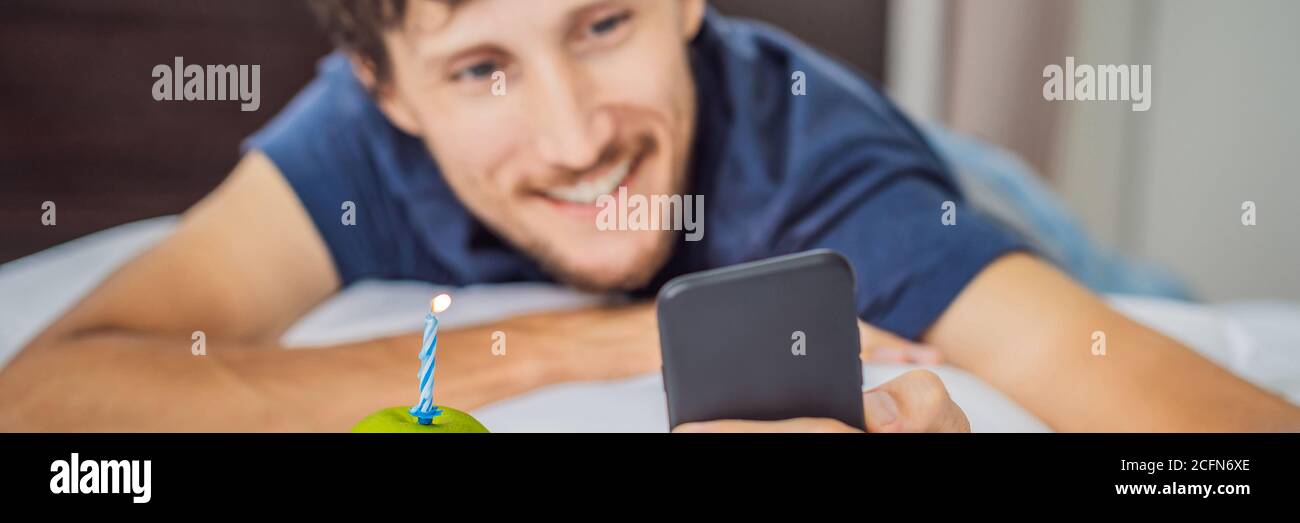 BANNER, LONG FORMAT Quarantine birthday. Man with a smartphone and birthday cake-apple with candle. Stay home while quarantine. Online call. Video Stock Photo