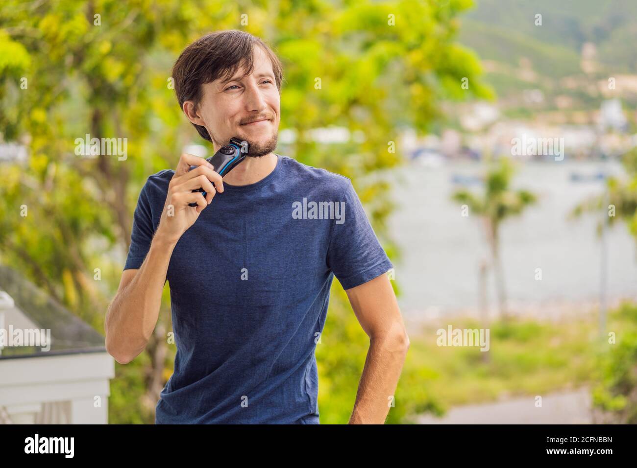 man is shaving off his beard with electric razor on the balcony during quarantine. Handsome bearded man trimming his beard with a trimmer at home Stock Photo