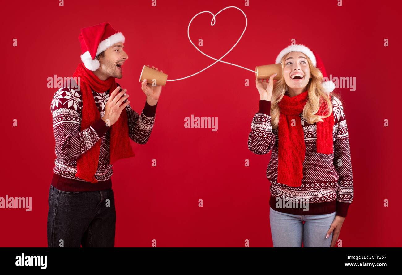 Christmas love message. Cute couple in Santa hats and sweaters talking on tin phone on red background Stock Photo