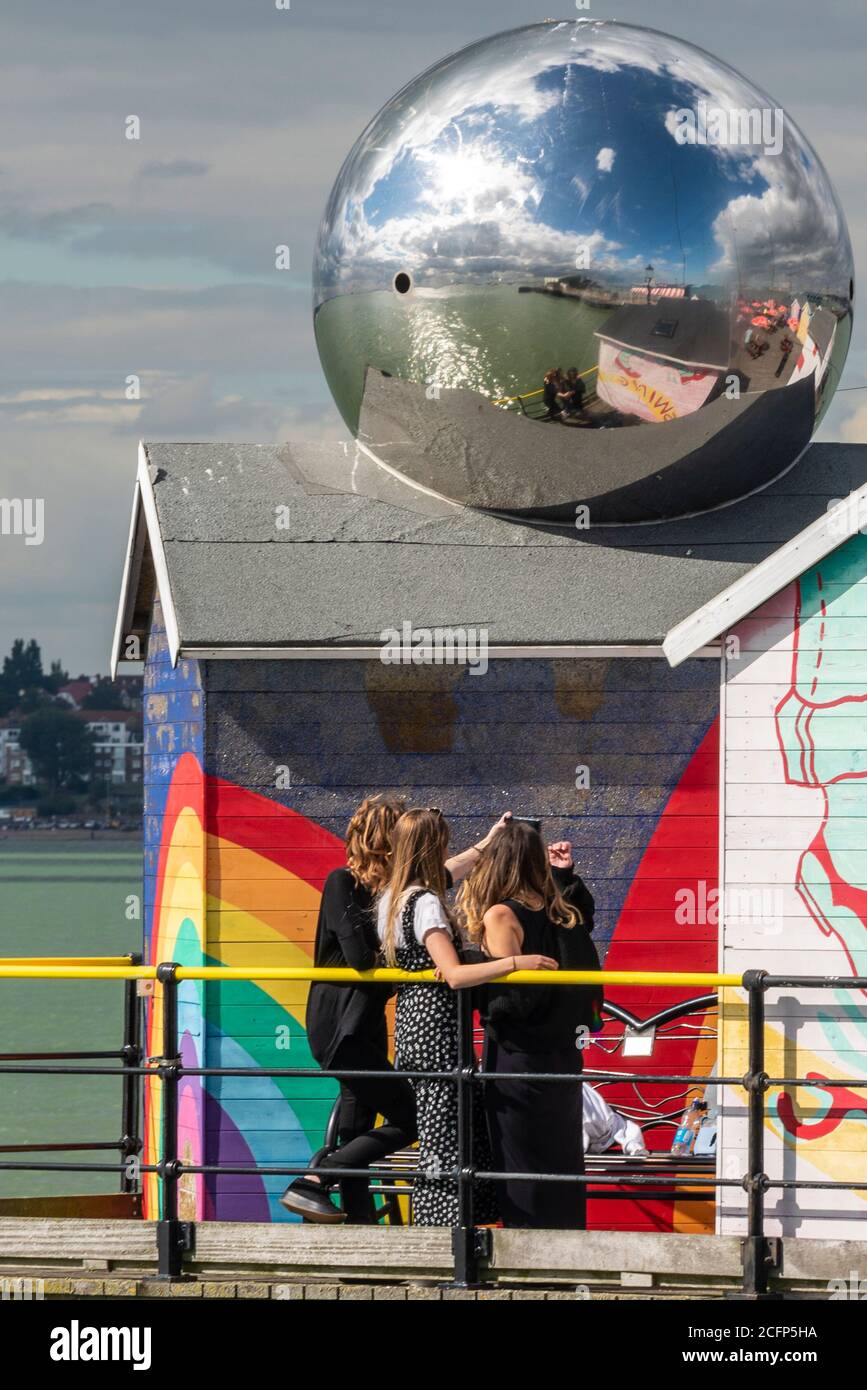 Three white Caucasian females taking a selfie photograph close together, reflected in large mirror ball. Reflecting friends. Brightly coloured huts Stock Photo