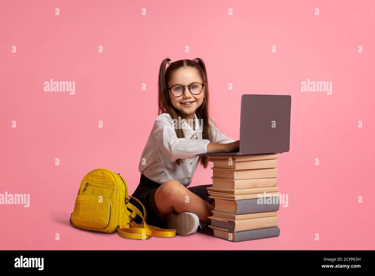 Smiling girl in glasses with backpack sits near stack of books and laptop Stock Photo