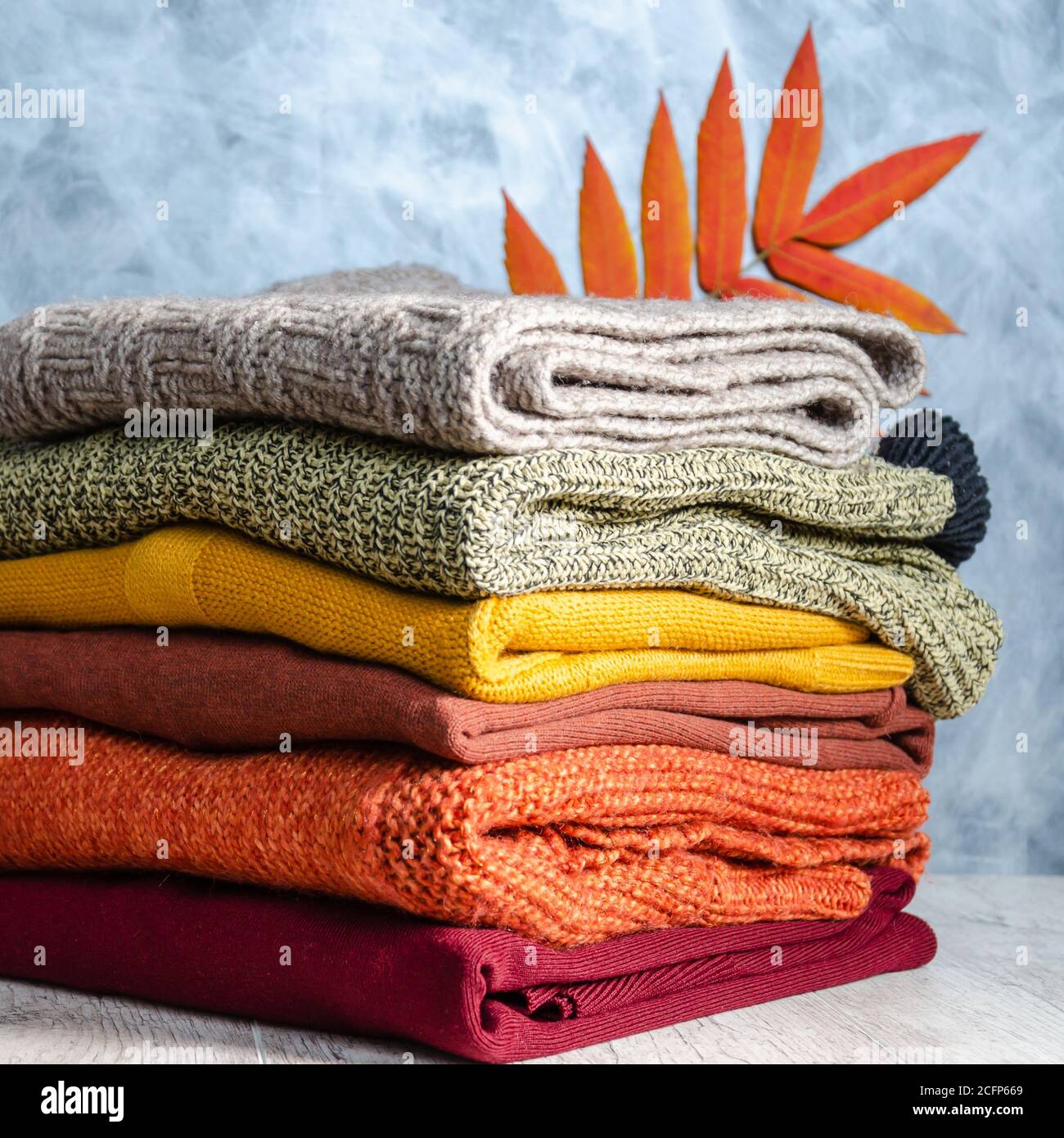 Stack of folded warm knitted women's sweaters in warm colors and bright autumn leaves on gray background. Copy space. Closeup Stock Photo