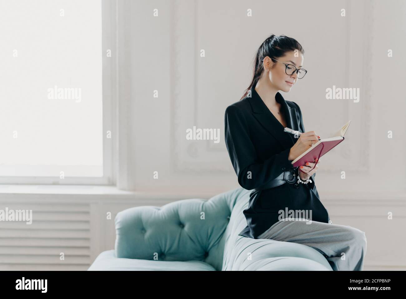 Attracative successful businesswoman in elegant clothes records information in notepad, sits on back of sofa, poses in home office, plans working proc Stock Photo