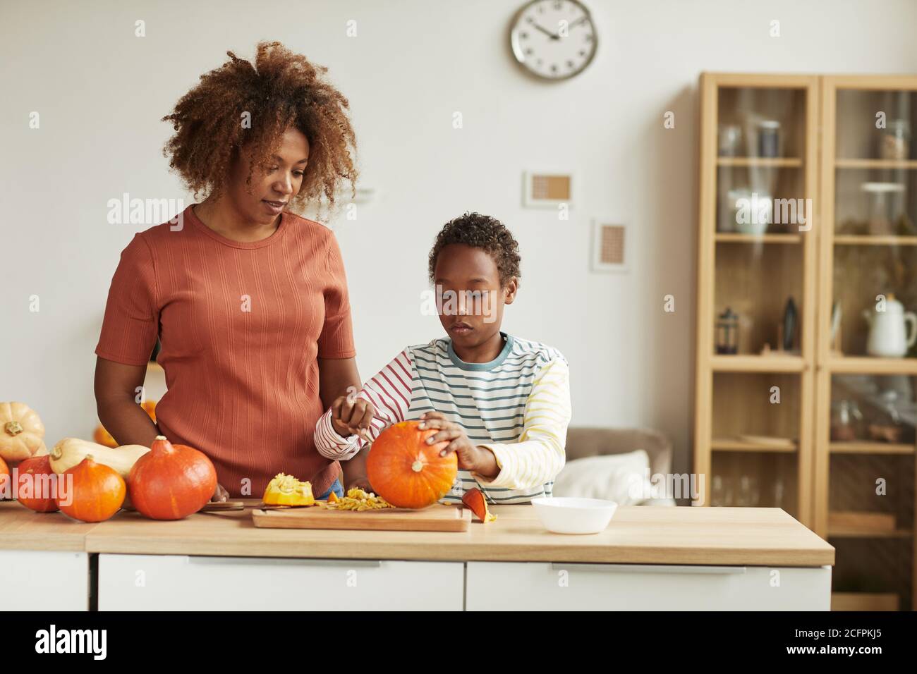 Young adult African American woman wearing casual outfit standing at table watching her child carving pumpkin Stock Photo