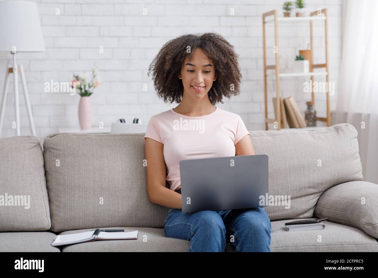 Friendly african american girl working in laptop, sitting on couch with ...