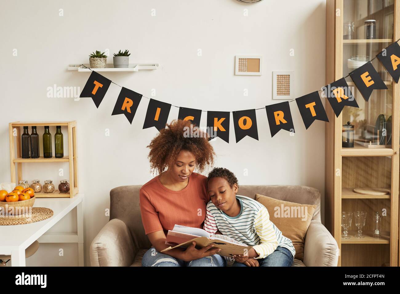 Modern African American mother sitting on sofa in living room with her young son reading spooky Halloween stories Stock Photo