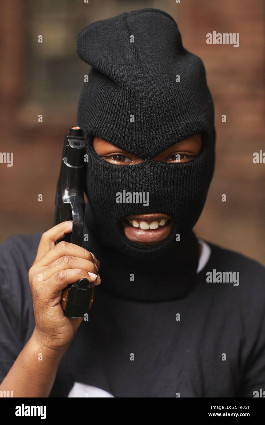Vertical close-up head and shoulders portrait of cool little boy dressed up as gangster for Halloween wearing balaclava holding toy gun smiling at camera Stock Photo