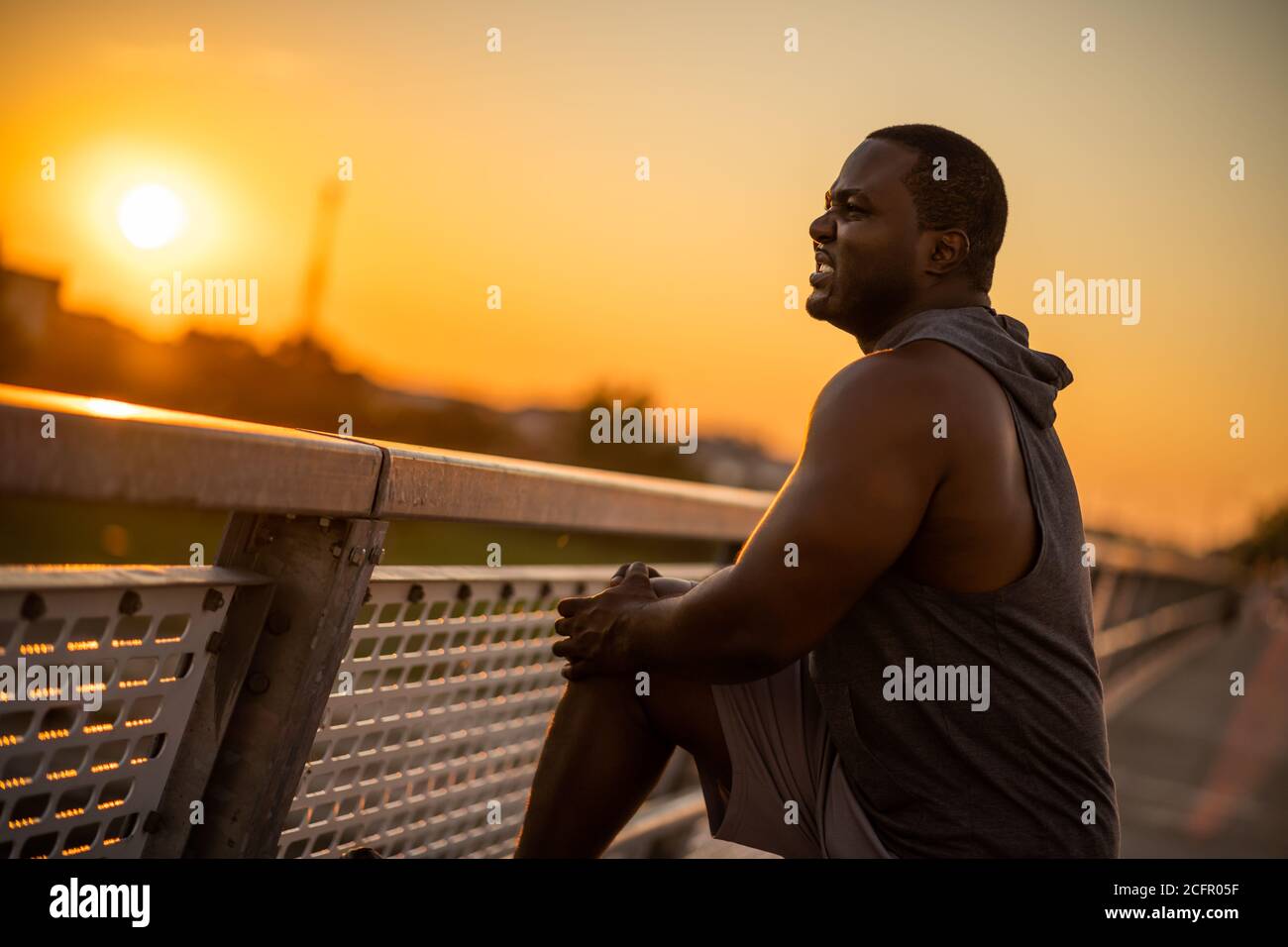 Young african-american man is having pain in knee while exercising in sunset on the bridge in the city. Stock Photo
