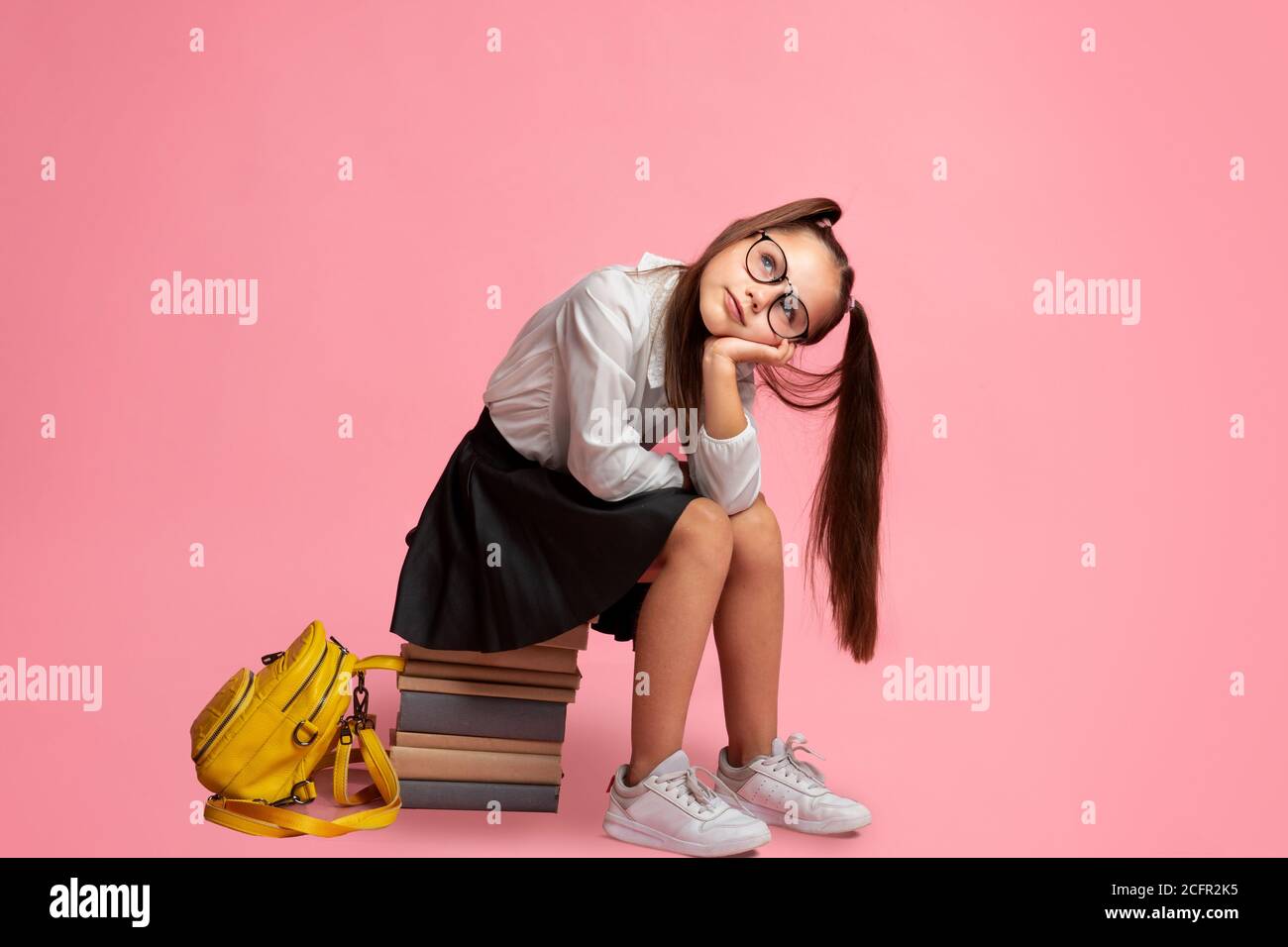 Schoolgirl in glasses with ponytails and backpack sits on stack of books and looks at empty space Stock Photo