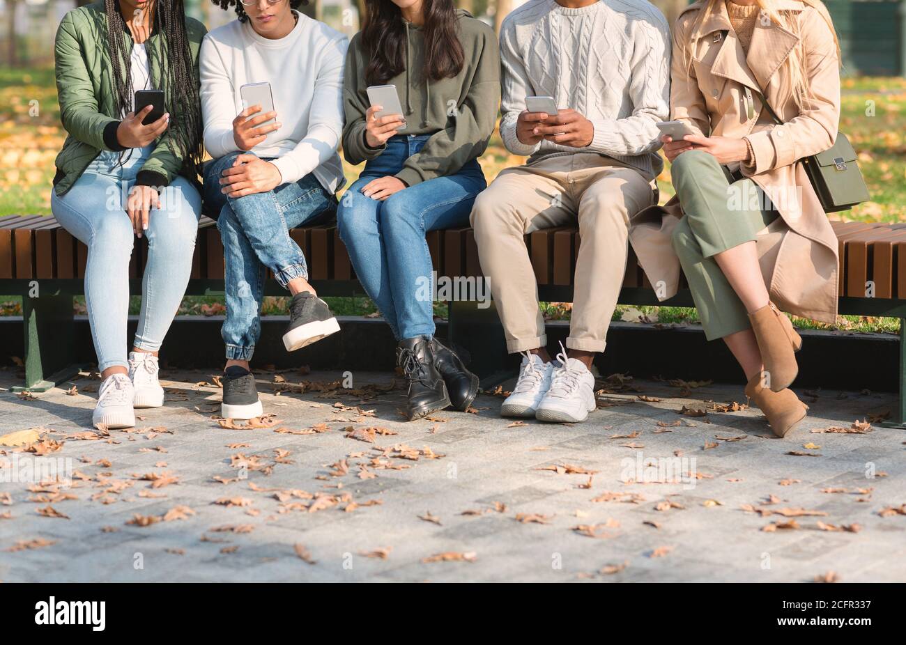 Cropped of young friends using mobile phones, sitting on bench Stock Photo