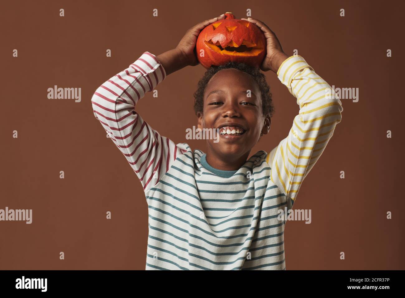 Cheerful African American boy wearing striped long-sleeve shirt holding Jack O' Lantern on his head smiling at camera, studio portrait Stock Photo