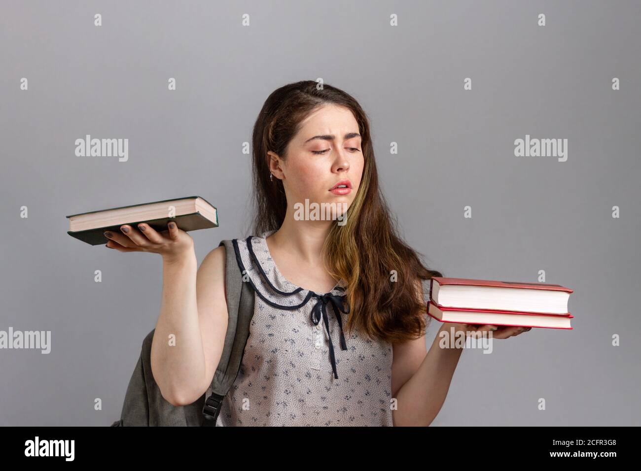 Education. A young woman or teenager with a backpack on her back, holding books in her hands, and looking at a large stack. Stock Photo