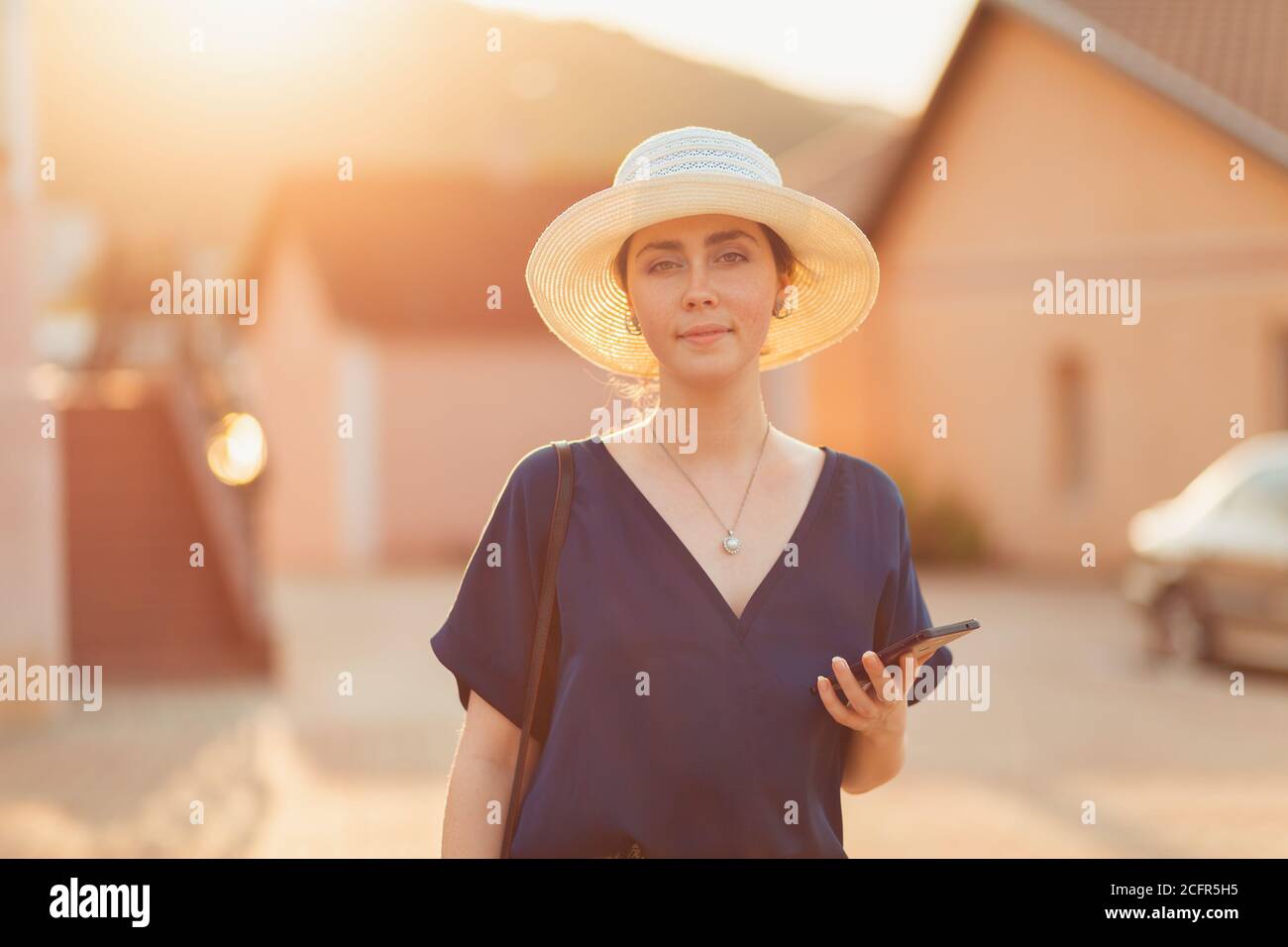 Communication. An elegant woman in a hat smiles and holds a smartphone in her hand. In the background of the house and sunset lighting in a blur. The Stock Photo