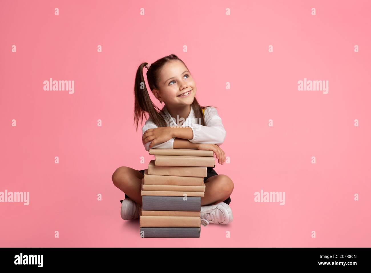 Cheerful schoolgirl dreaming, sitting at book stack and, looks at empty space Stock Photo