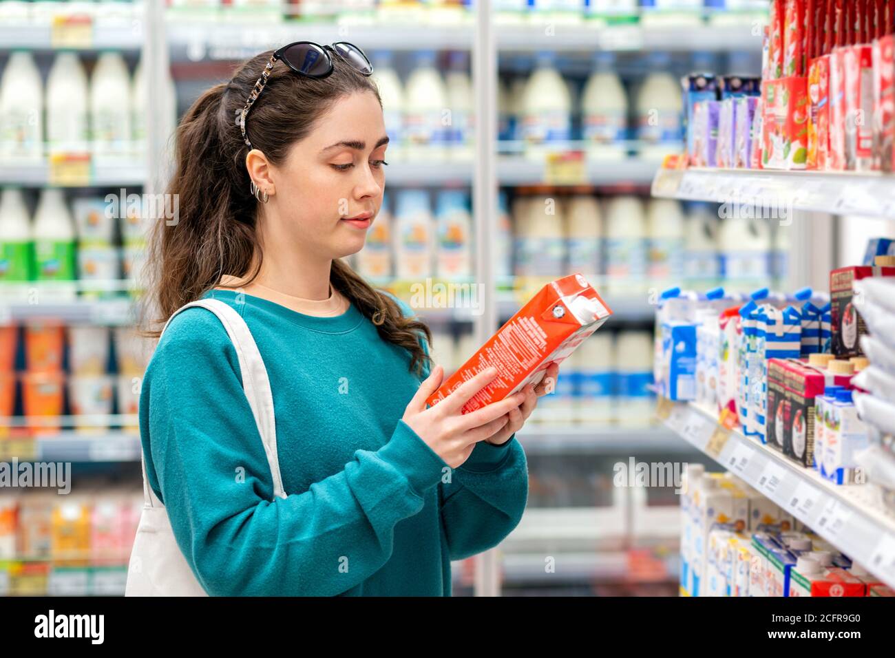 A young beautiful Caucasian woman with an eco-bag on her shoulder, reads the ingredients on the package. Concept of food purchase and shopping. Stock Photo